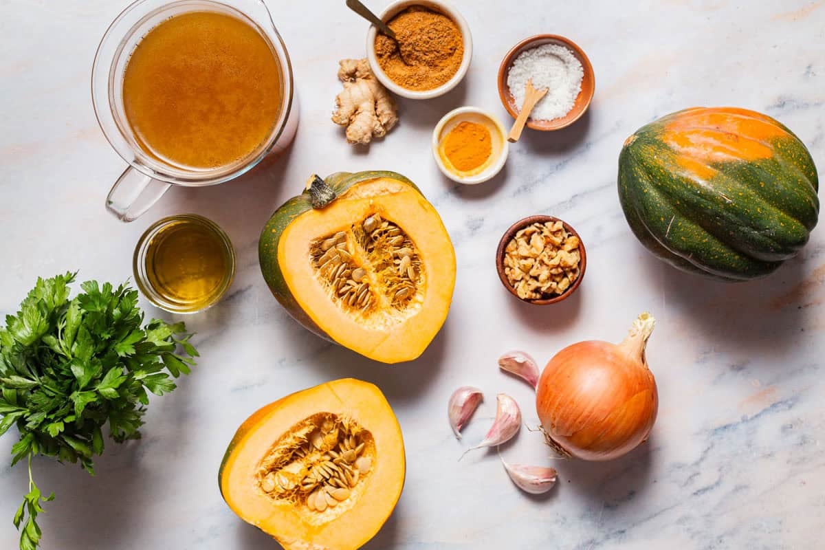 Ingredients for acorn squash soup including acorn squash, olive oil, onion, garlic, ras el hanout, turmeric, salt, vegetable broth, ginger, walnuts, and parsley.