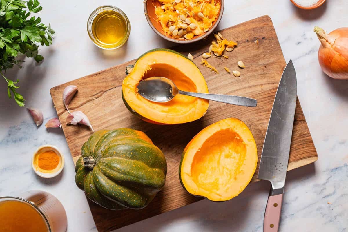 An overhead photo of 2 acorn squash have with their seeds removed on a cutting board, one with a spoon in it. Next to these is a knife, a whole acorn squash, vegetable broth, cloves of garlic, an onion, some parsley a bowl of the acorn squash seeds, and small bowls of salt, olive oil and turmeric.