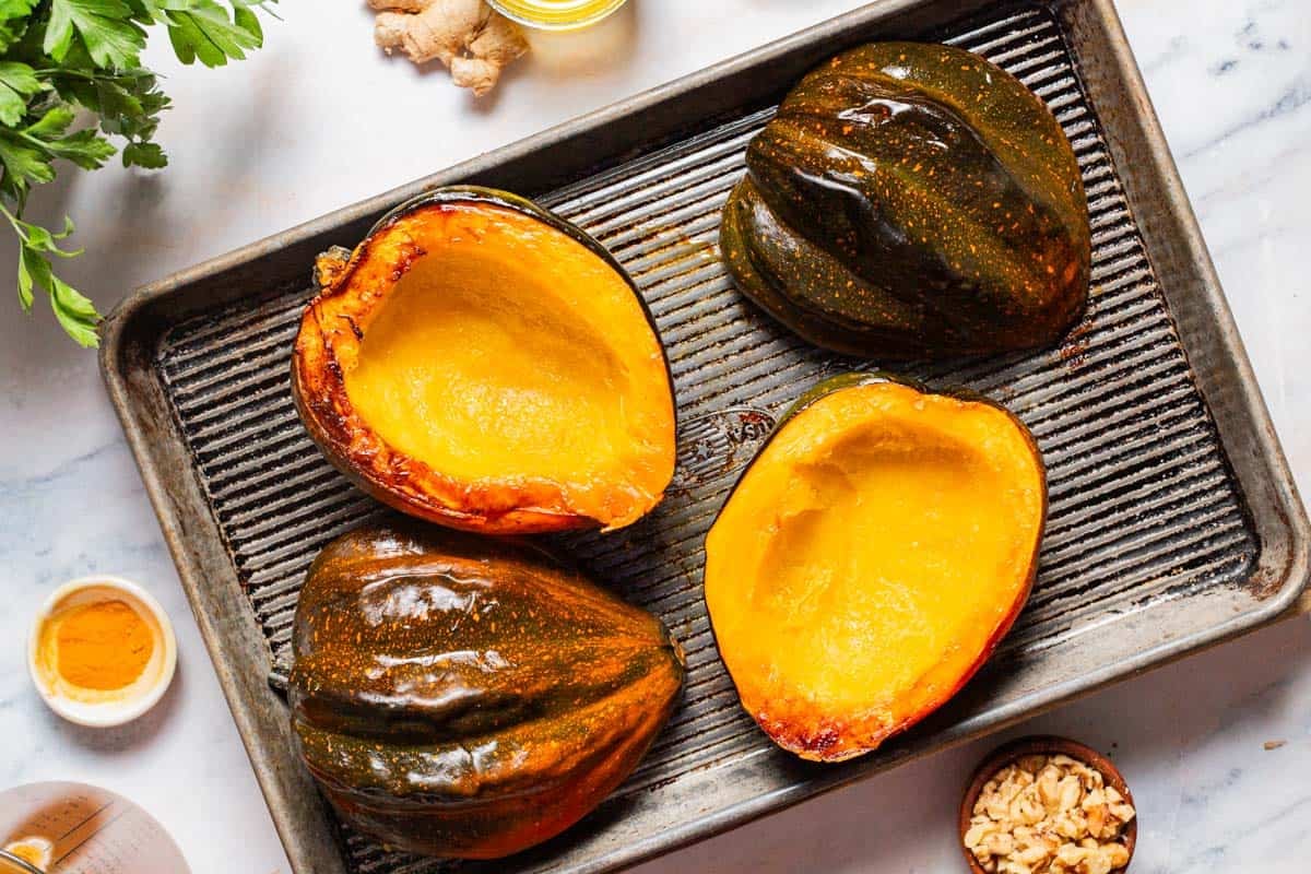 An overhead photo of 4 roasted acorn squash halves on a baking sheet. Next to this is some parsley, small bowls of walnuts and turmeric, and a bit of ginger root.