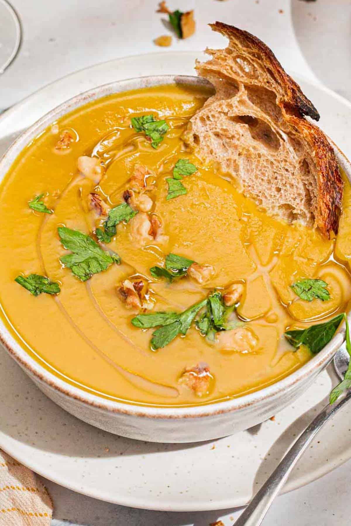 A close up of acorn squash soup garnished with walnuts, parsley and a drizzle of olive oil in a bowl with a piece of bread. Next to this is a spoon.