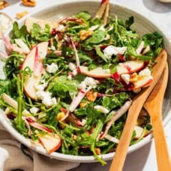 Apple walnut salad in a serving bowl with wooden serving utensils surrounded by bowls of salt and pepper and a cloth napkin.