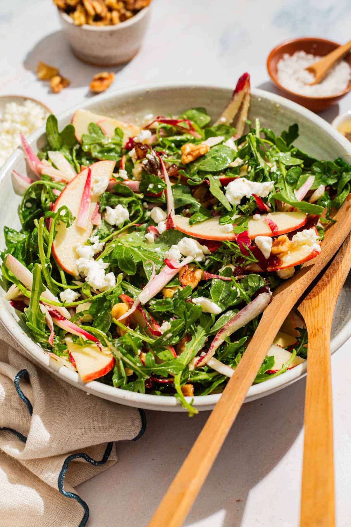 Apple walnut salad in a serving bowl with wooden serving utensils surrounded by bowls of salt and walnuts, and a cloth napkin.