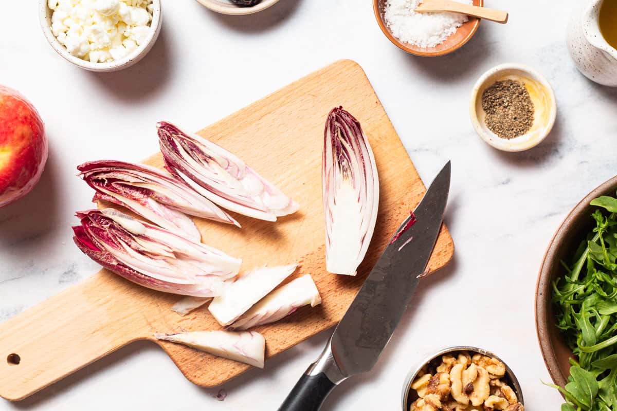 Sliced endives on a cutting board with a knife surrounded by an apple, apple cider vinaigrette, and bowls of goat cheese, salt, pepper, walnuts and arugula.