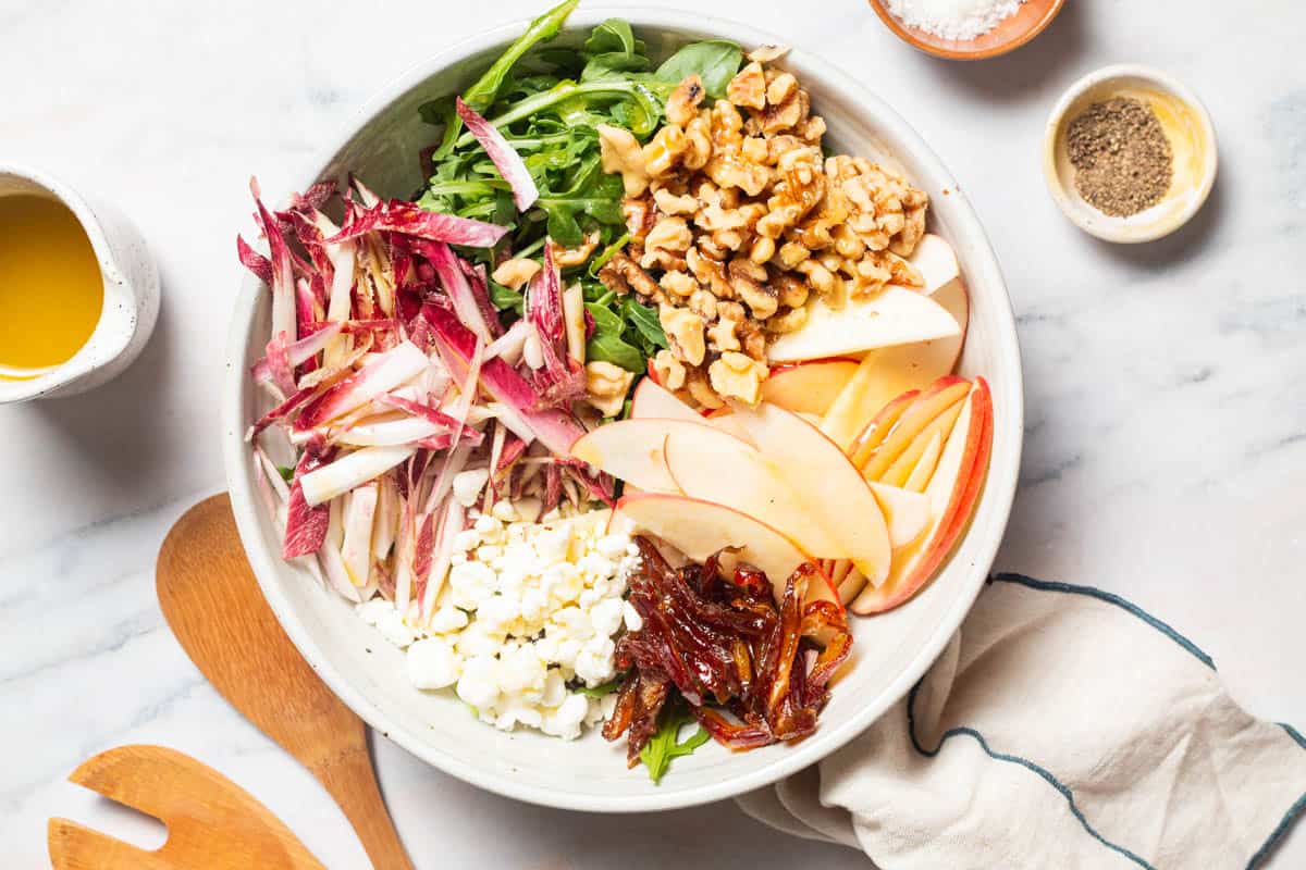 An overhead photo of the ingredients for the apple walnut salad in a serving bowl before being mixed together. This is surrounded by bowls of salt and pepper, wooden serving utensils, apple cider vinaigrette, and a cloth napkin.