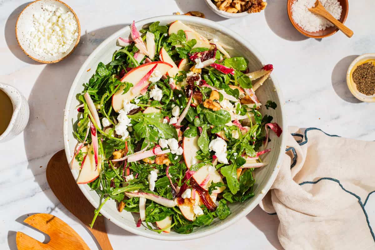 An overhead photo of apple walnut salad in a serving bowl surrounded by bowls of crumbled goat cheese, salt, pepper and walnuts, wooden serving utensils, apple cider vinaigrette, and a cloth napkin.