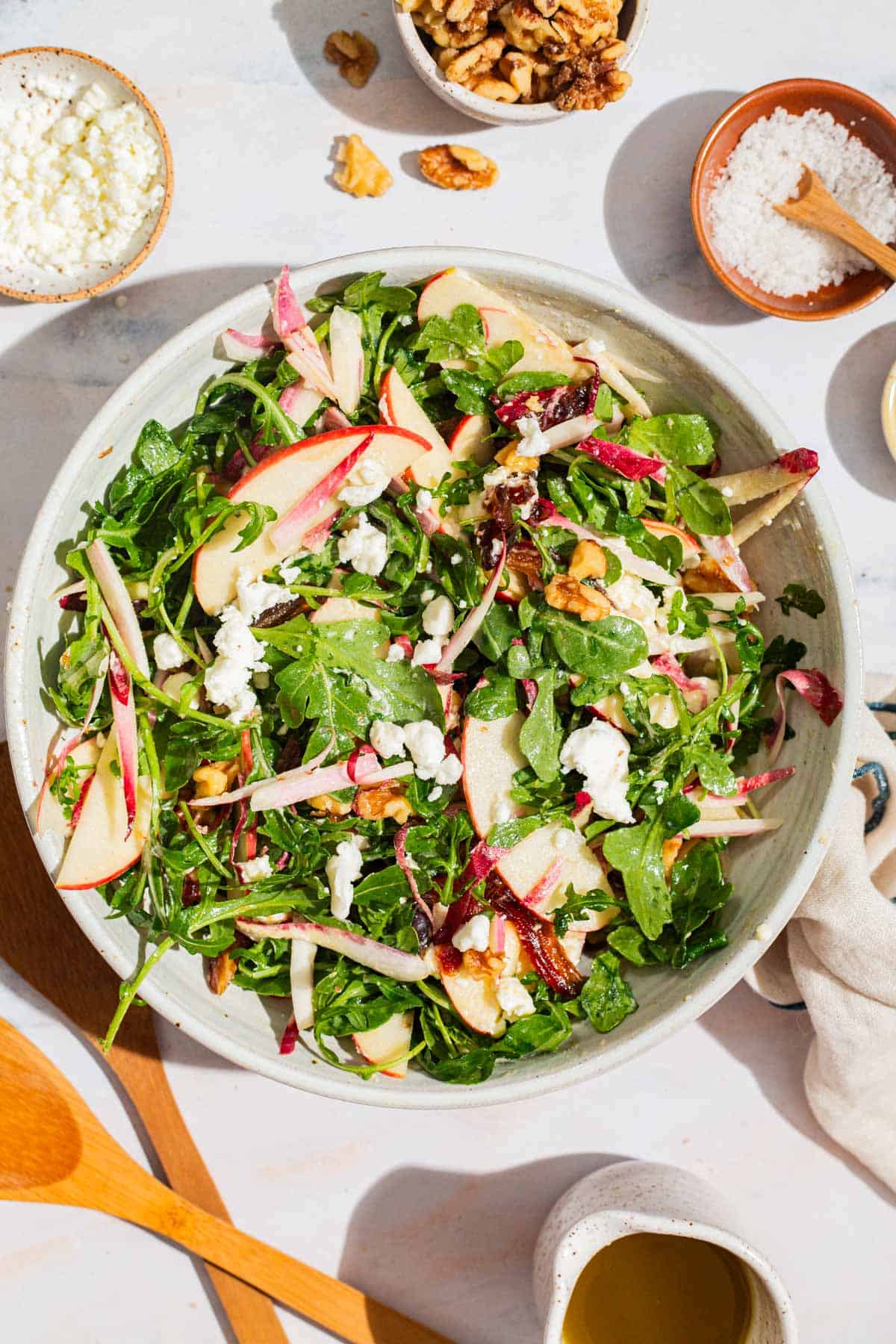 An overhead photo of apple walnut salad in a serving bowl surrounded by bowls of crumbled goat cheese, salt, and walnuts, wooden serving utensils, apple cider vinaigrette, and a cloth napkin.