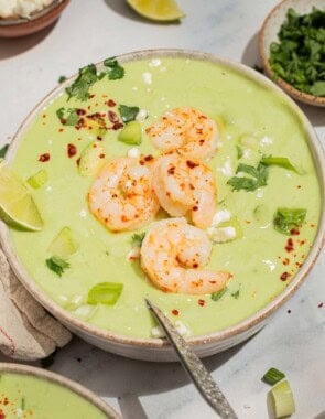 A close up of avocado soup topped with shrimp, cilantro, feta, aleppo pepper and scallions in a bowl with a spoon. Next to this is a lime wedge and bowls of feta and cilantro.