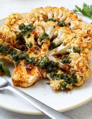 Side shot of a cauliflower steak on a white plate with a metal fork.