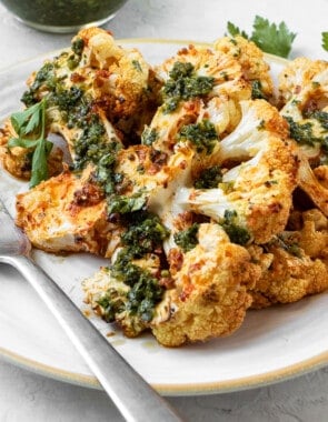 Two cauliflower steaks attractively layered on a plate with one resting on top of the other. Both have been dressed with green Chermoula. There is a small bowl of Chermoula on the side and a metal fork on the plate.