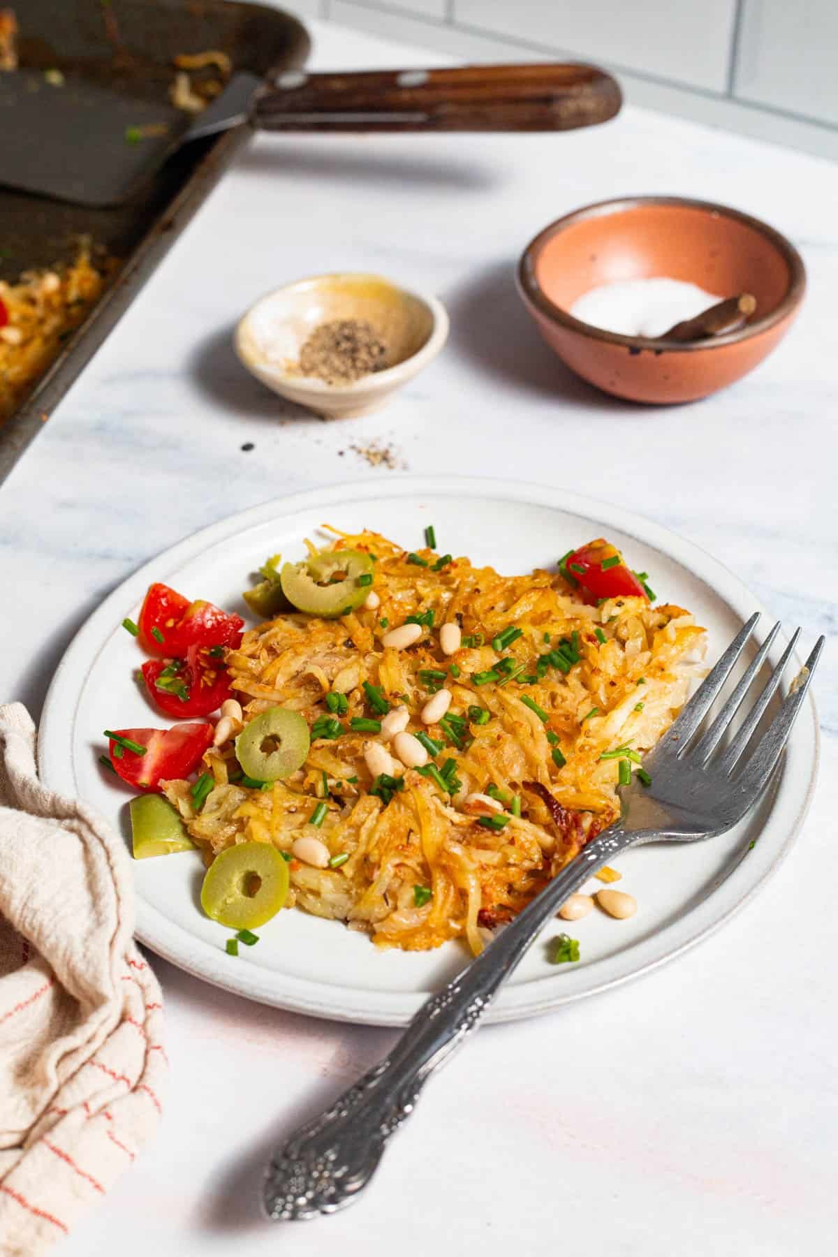 A serving of baked hash browns on a plate with a fork. Surrounding this is a kitchen towel, the sheet pan with a spatula, and bowls of salt and pepper.