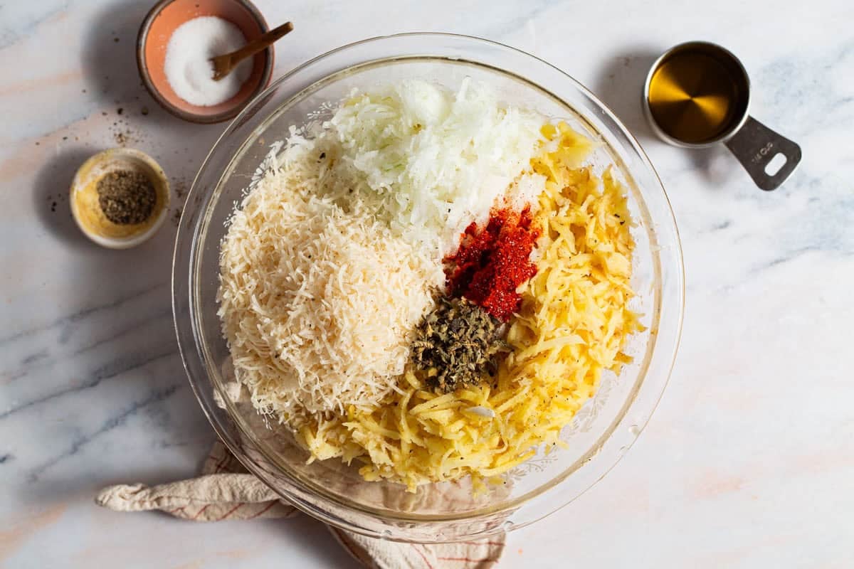 The ingredients for baked hash browns in a bowl just before being mixed together. Surrounding this is a 1/4 cup of olive oil, bowls of salt and pepper, and a kitchen towel.