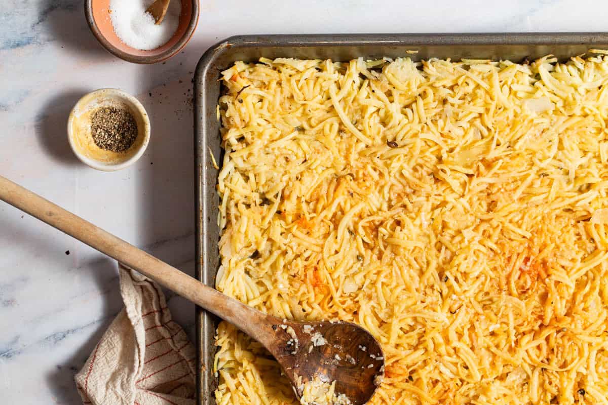 The hash brown mixtures spread evenly on a sheet pan with a wooden spoon. Next to this is a kitchen towel and bowls of salt and pepper.