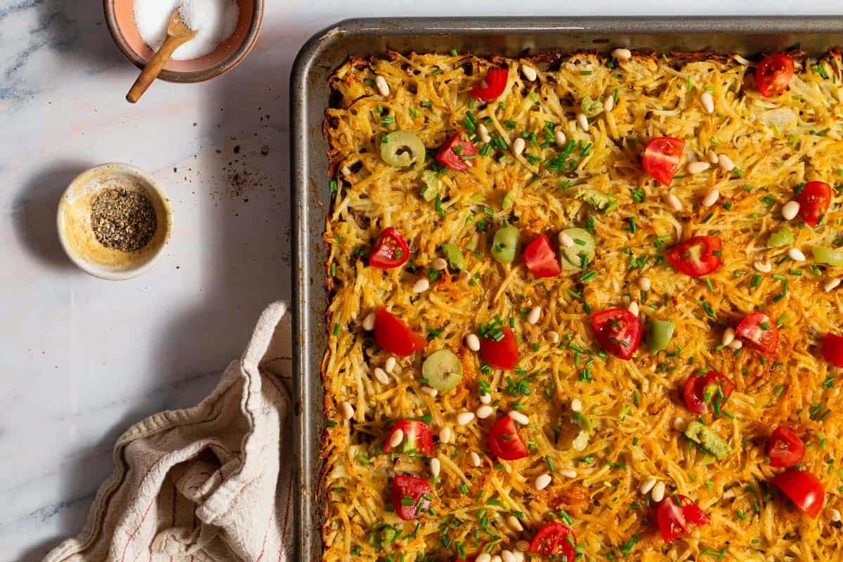 Baked hash browns on a sheet pan surrounded by a kitchen towel and bowls of salt and pepper.