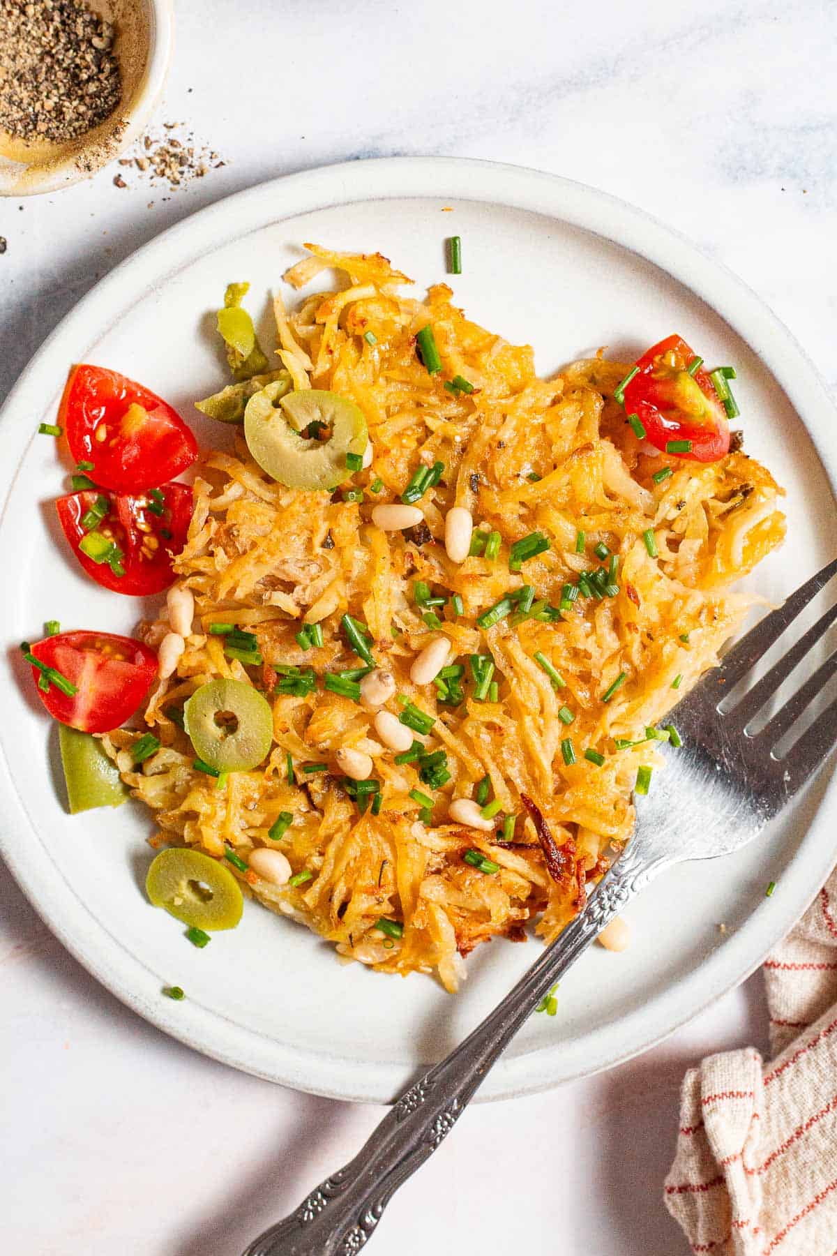 An overhead photo of a serving of baked hash browns on a plate with a fork. Next to this is a kitchen towel and a bowl of pepper.