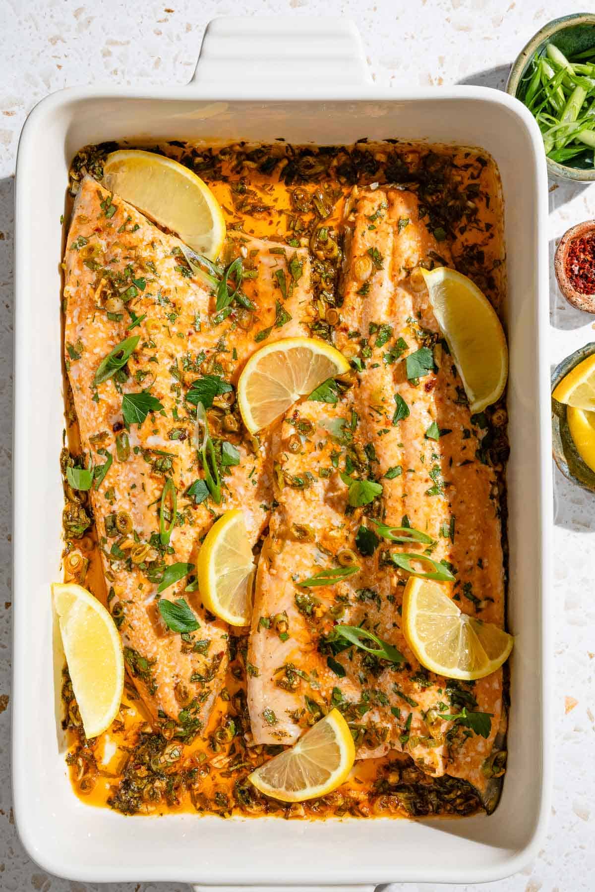 An overhead photo of baked trout garnished with parsley and green onions in a baking dish. This is surrounded by small bowls of green onions, urfa biber pepper and lemon wedges.