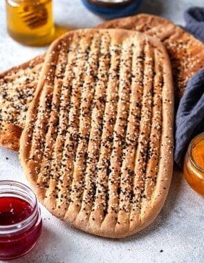 two pieces of barbari bread surrounded by bowls of toppings such as feta cheese, honey, and jam.