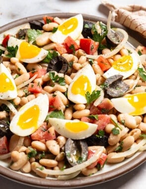 A close up of cannellini bean salad on a serving platter with a fork. Next to this is a bowl of salt and a cloth napkin.