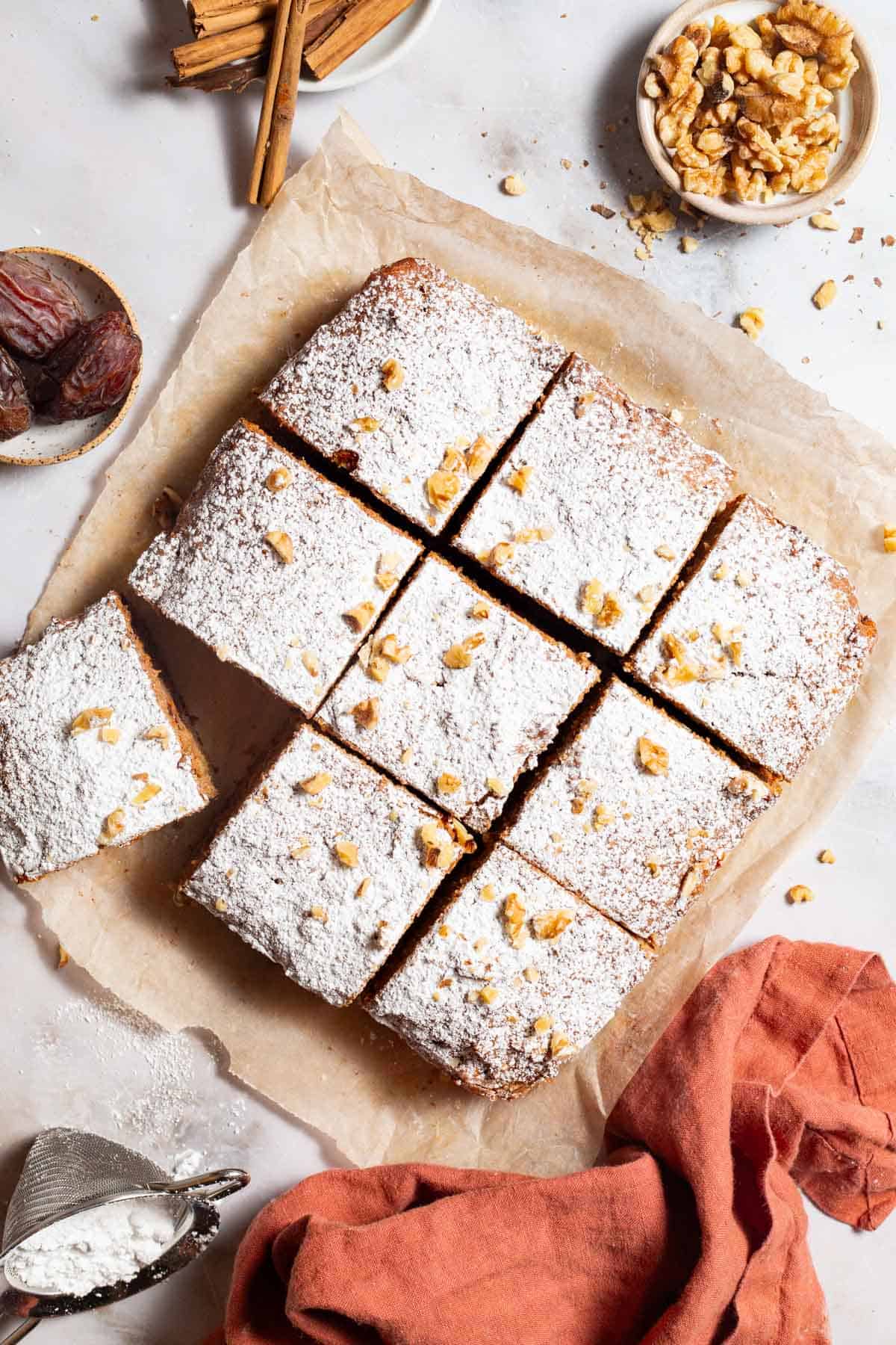 An and overhead photo of 9 slices of healthy carrot cake on a sheet of parchment paper. Next to these are bowls of walnuts, cinnamon sticks and dates, a sieve with powdered sugar, and a kitchen towel.