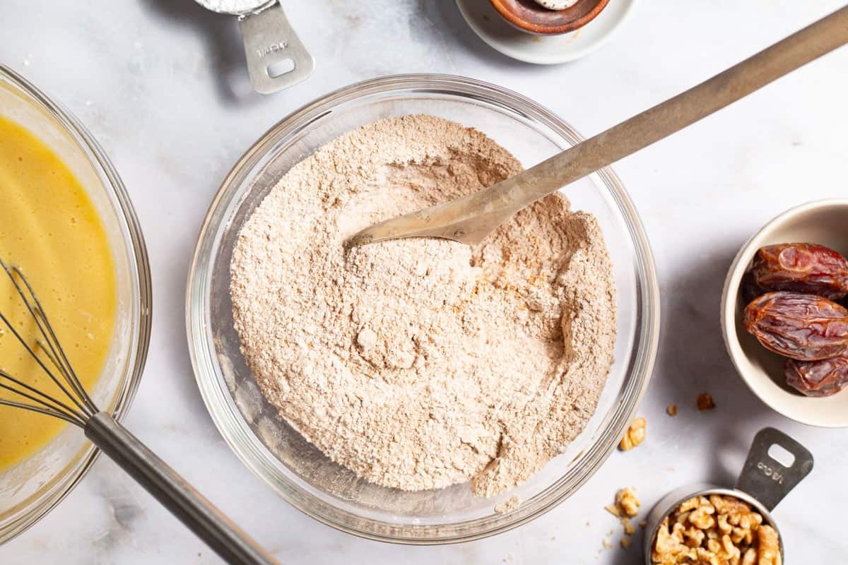 A bowl of the dry ingredients for the healthy carrot cake with a wooden spoon. Next to this is the bowl with the wet ingredients and a whisk, a measuring cup with walnuts and a bowl of dates.