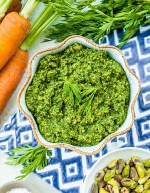 Carrot top pesto in a bowl next to a bunch of carrots and bowls of salt and pistachios.