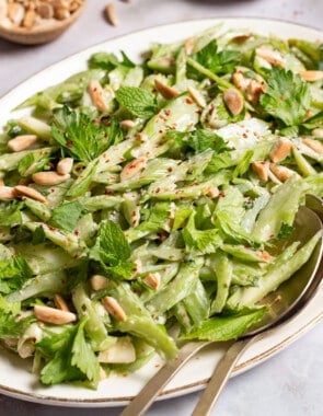 Celery salad on a serving platter with serving utensils. Next to this are bowls of slivered almonds, black pepper, salt and a lemon half.