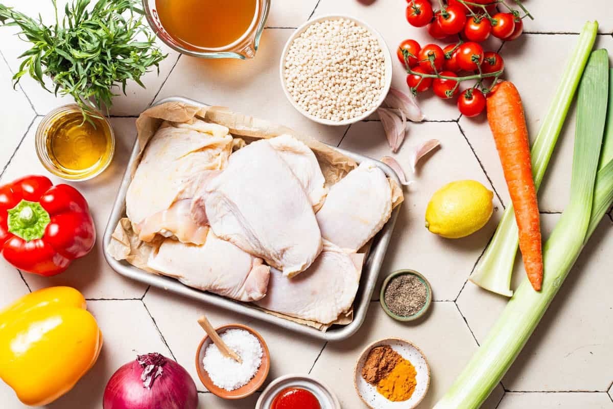 Ingredients for one pan tomato chicken with baharat, tarragon, and couscous including chicken thighs, bell peppers, a red onion, celery, a carrot, a leek, cherry tomatoes, garlic, olive oil, salt, black pepper, baharat, turmeric, tomato paste, pearl couscous, broth, lemon and taragon.