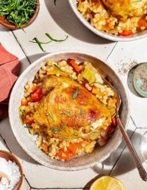 An overhead photo of a serving of the one pan tomato chicken with baharat, tarragon, and couscous in a bowl with a fork. Next to this is a lemon half, small bowls of salt, pepper and tarragon, and a bowl with another serving of the chicken.