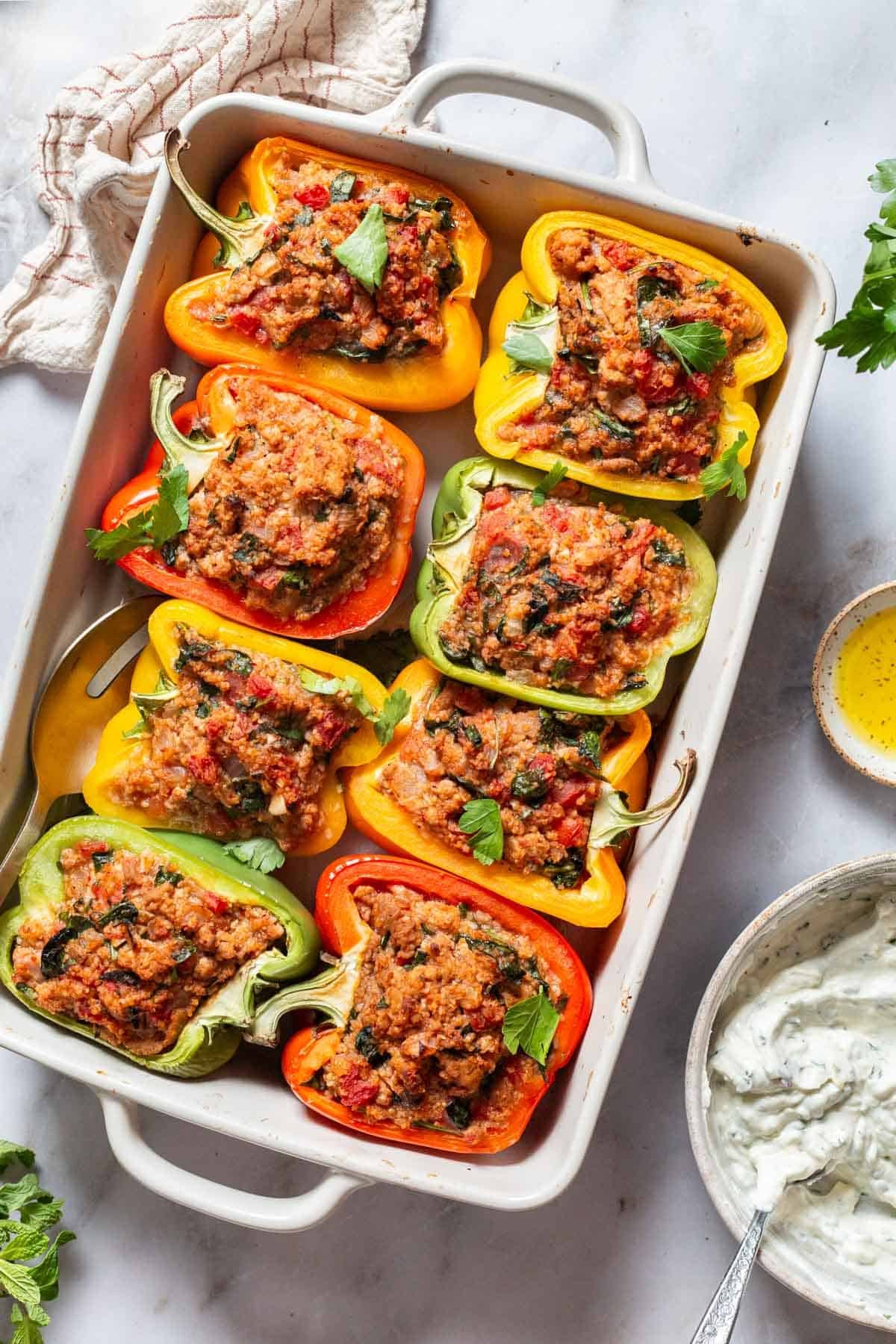 An overhead photo of 8 cooked chicken stuffed peppers in a baking dish with a serving spoon. Surrounding this is the yogurt sauce in a bowl with a spoon, a small bowl of olive oil, and a cloth napkin.