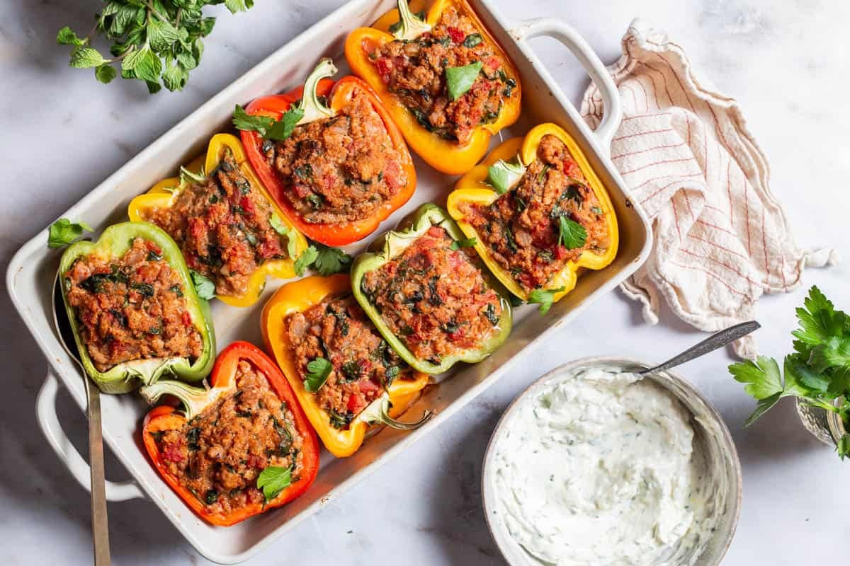 An overhead photo of 8 cooked chicken stuffed peppers in a baking dish with a serving spoon. Surrounding this is a bunch of mint, a bunch of parsley, yogurt sauce in a bowl with a spoon, and a cloth napkin.