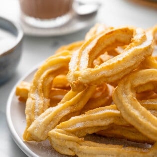 Stack of churros on a gray plate with Spanish drinking chocolate in the background.