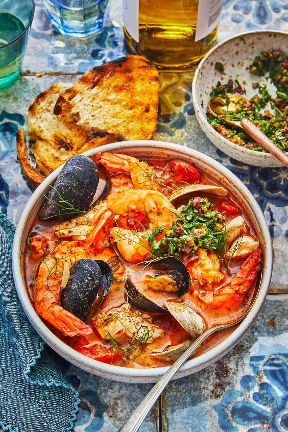 a bowl of cioppino with a spoon, two pieces of toasted and a bowl of Parsley-Olive Gremolata.