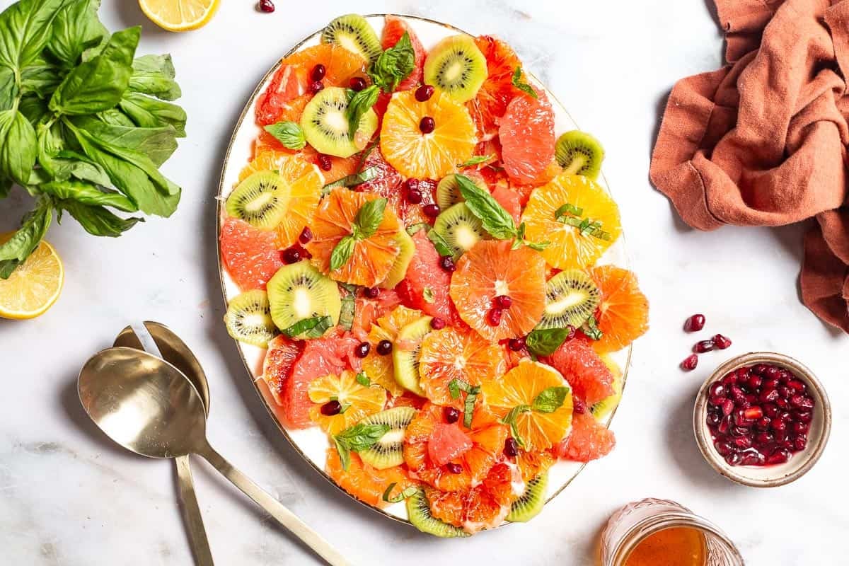 an overhead photo of citrus salad on a serving platter surrounded by a cloth napkin, gold serving utensils, basil, lemons, a small bowl of pomegranate seeds and a jar of dressing.