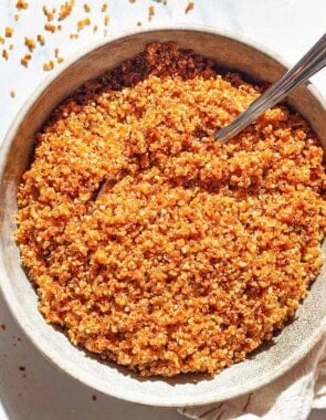 An overhead photo of crispy quinoa in a bowl with a spoon. Next to this is a kitchen towel and bowls of aleppo, olive oil, and kosher salt.