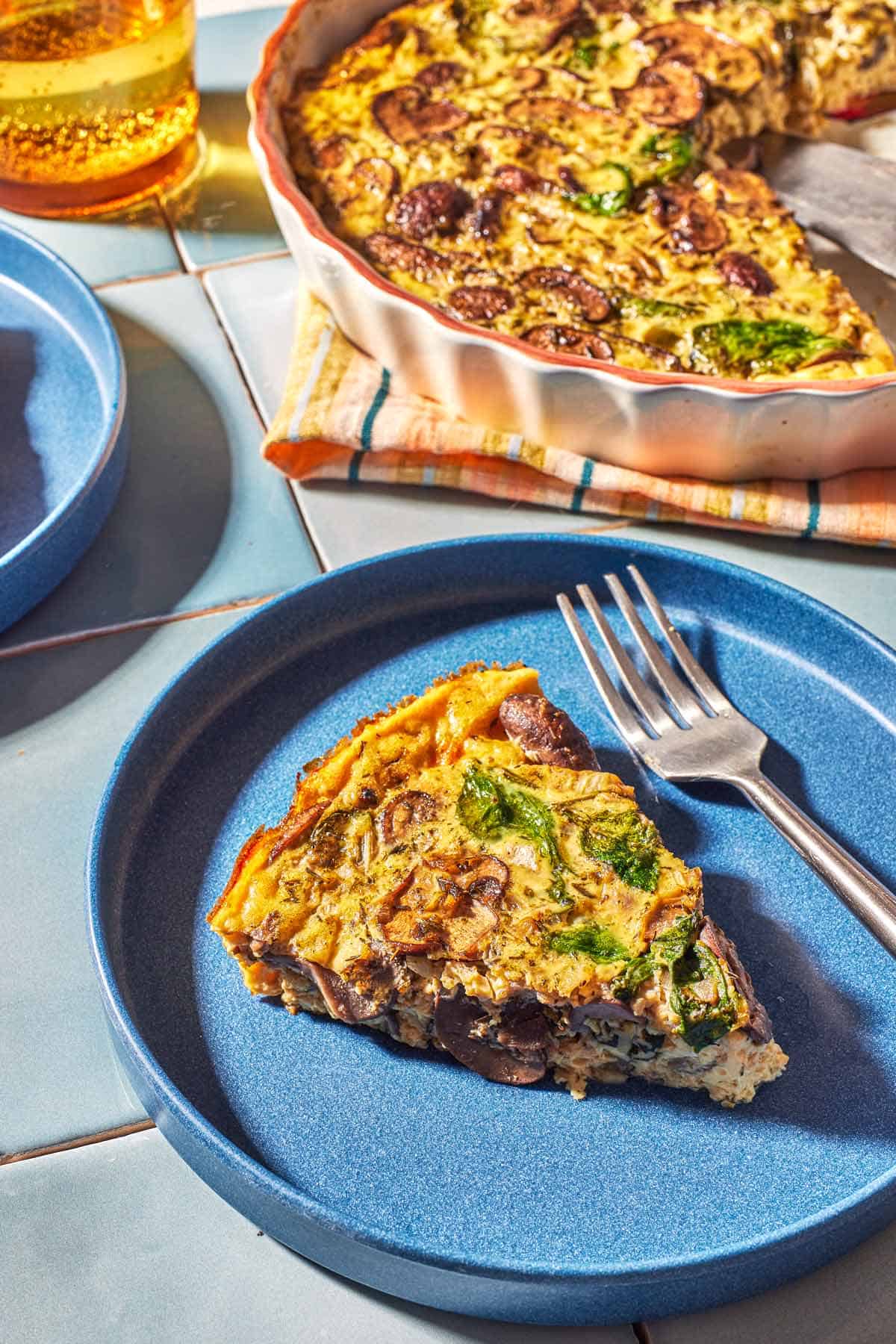 Slice of crustless quiche with spinach and mushrooms on a blue plate. The rest of the quiche is in the background. 