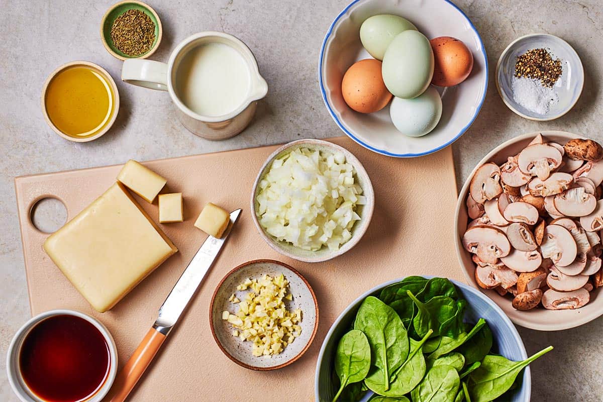 ingredients for crustless quiche including eggs, milk, fontina cheese, olive oil, baby spinach, sliced mushrooms, onion, garlic, thyme, salt, pepper and balsamic vinegar.