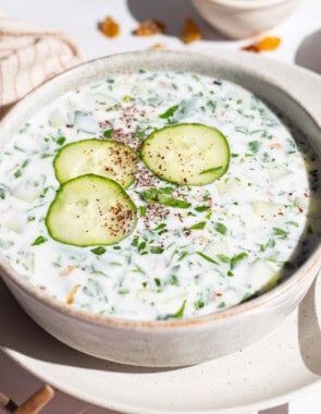 A bowl of cucumber soup garnished with sumac, parsley and cucumber slices on a plate with a spoon. Next to this is a kitchen towel and bowls of sumac and walnuts.