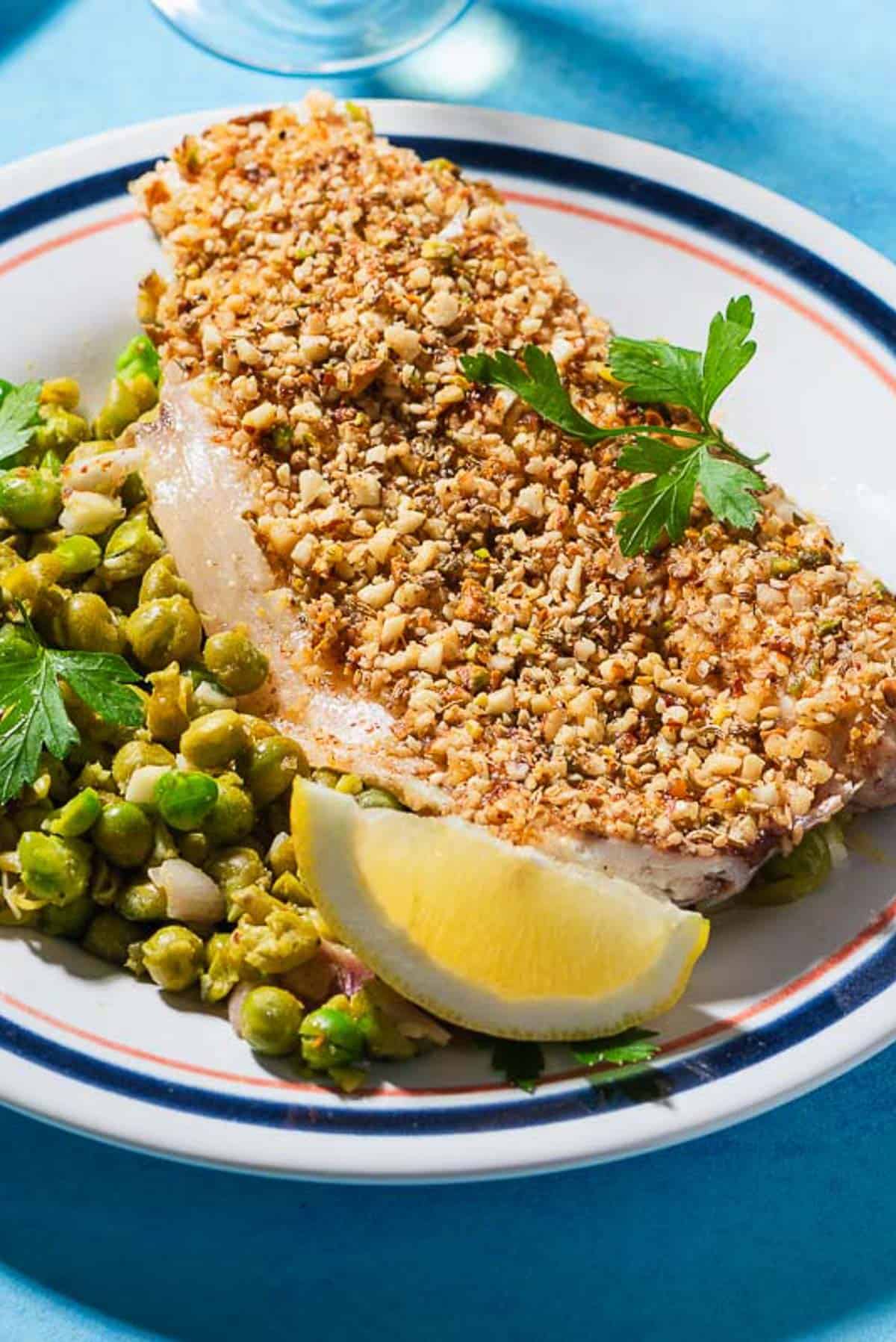 A close up of a serving of baked white fish topped with dukkah on a bed of smashed peas on a plate with a lemon wedge.