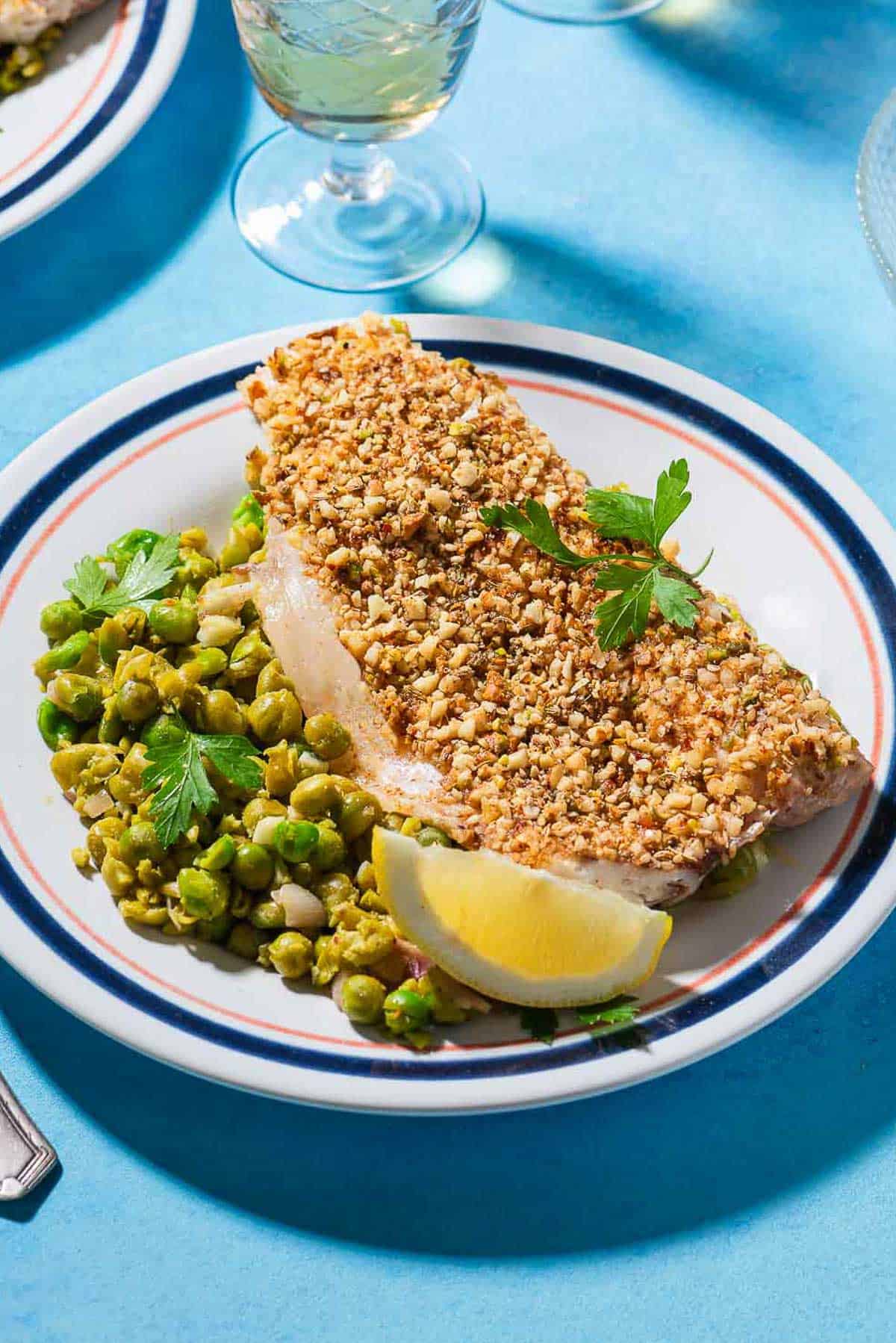 A close up of a serving of baked white fish topped with dukkah on a bed of smashed peas on a plate with a lemon wedge. Behind this is a glass of wine.