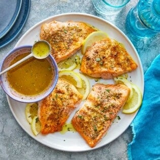 four broiled salmon fillets on a plate with lemon slices and a bowl of greek ladolemono dressing with a spoon.