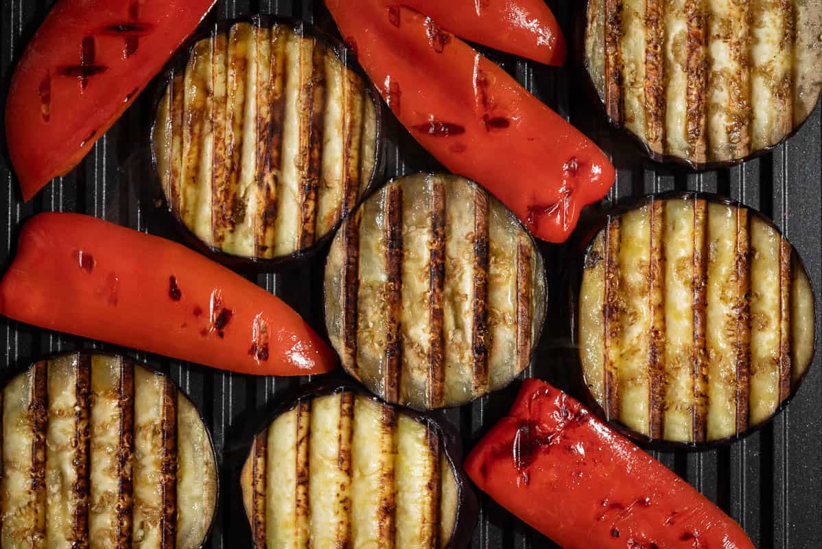 An overhead photo of eggplant and red bell pepper slices being grilled on a griddle.