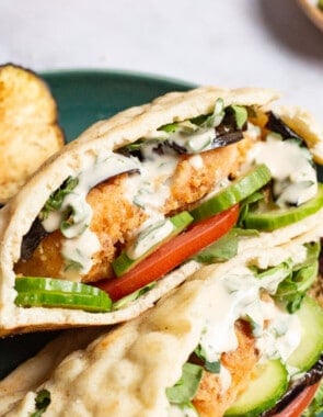 A close up of two fried fish sandwiches on a plate with sides of roasted eggplant slices, tomato, and a lemon wedge.