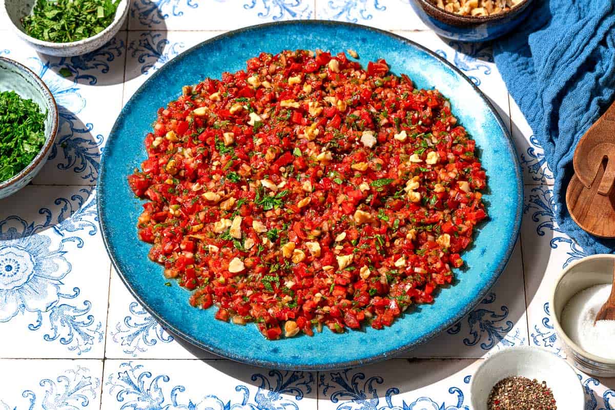 Ezme turkish salsa on a platter. Next to this are bowls of mint, parsley, walnuts, spices, salt, as well as wooden serving utensils on a cloth napkin.