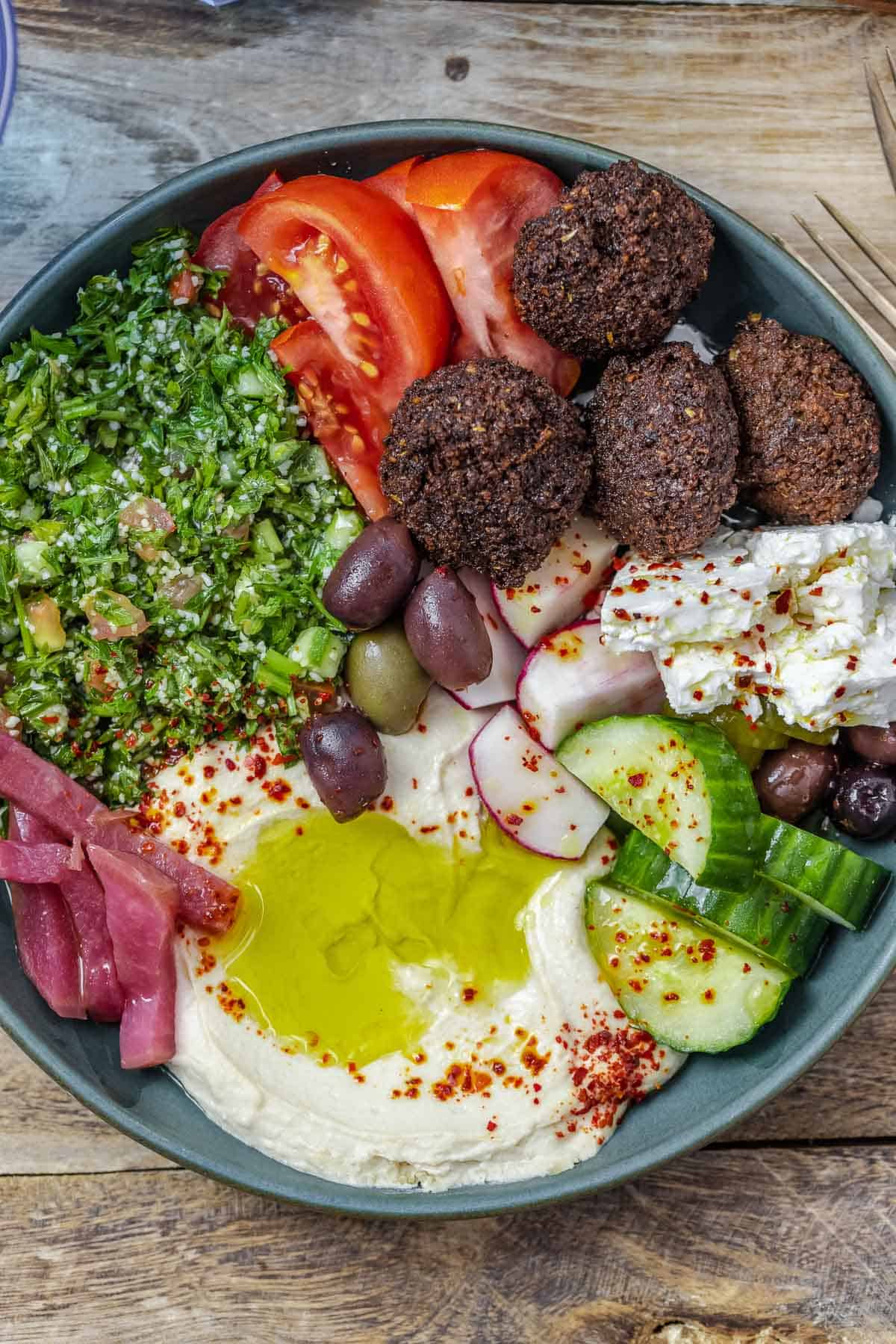 a close up of a falafel bowl assembled with hummus, pickles, olives , fresh vegetables and a tabouli salad