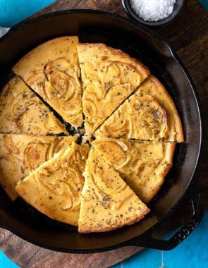 a baked farinata italian chickpea pancake sliced into pieces in a cast iron skillet next to a bowl of flaky sea salt.