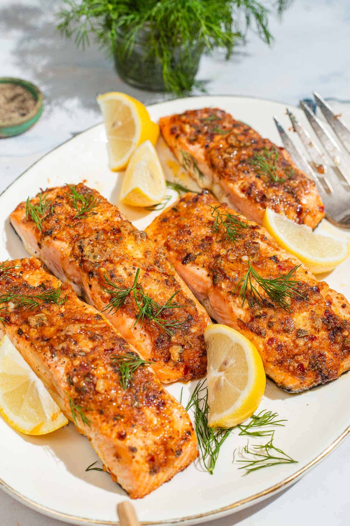 A close up of 4 baked dijon salmon fillets on a serving platter with lemon wedges and a fork. Next to this is a bowl of black pepper and a bunch of dill.