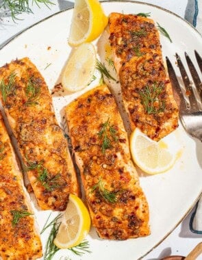 An overhead photo of 4 baked dijon salmon fillets on a serving platter with lemon wedges and a fork. Next to this is a kitchen towel and a plate of lemon wedges.