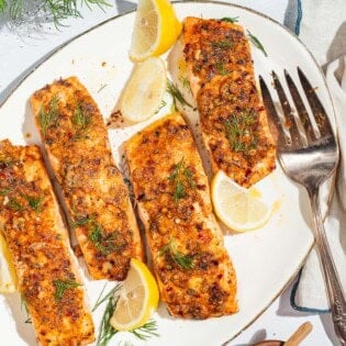 An overhead photo of 4 baked dijon salmon fillets on a serving platter with lemon wedges and a fork. Next to this is a kitchen towel and a plate of lemon wedges.