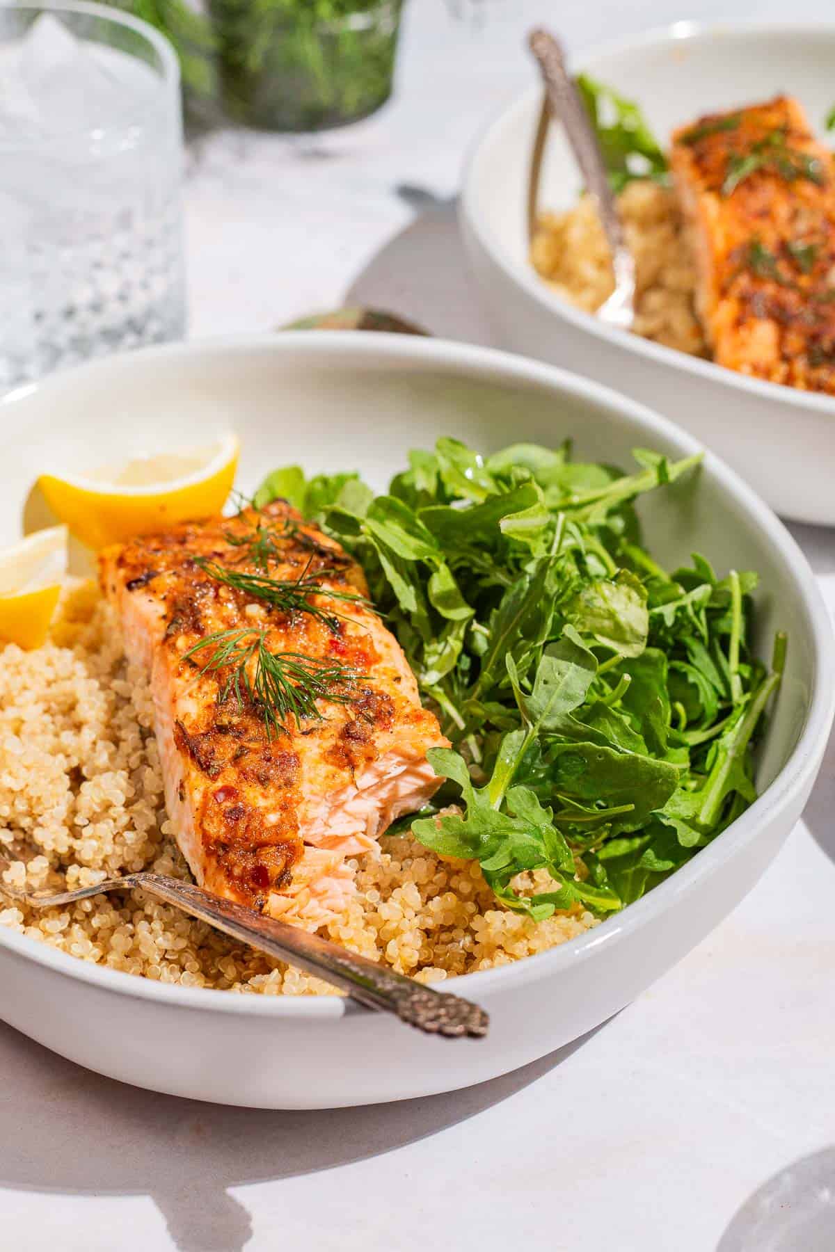 A close up of a serving of baked dijon salmon in a bowl with quinoa, arugula, lemon wedges and a fork. Behind this is another serving of the salmon in a bowl, and a glass of water.