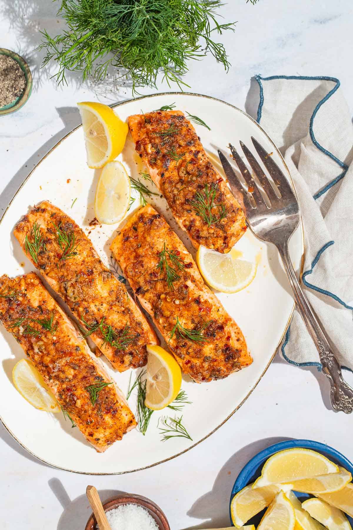 An overhead photo of 4 baked dijon salmon fillets on a serving platter with lemon wedges and a fork. Next to this is a kitchen towel, some dill, bowls of salt and pepper, and a plate of lemon wedges.