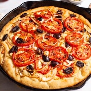 A close up of greek pizza in a cast iron skillet next to small bowls of kalamata olives and salt, and a kitchen towel.